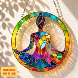 May include: A stained glass suncatcher featuring a seated figure in a yoga pose, within a circular frame. The figure and frame are composed of colorful glass pieces in shades of red, blue, yellow, and green. The suncatcher hangs from a silver chain.