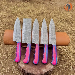 May include: A set of five Damascus steel kitchen knives with pink and blue handles, resting on a wooden stump. The blades have a unique patterned design, and the handles are a vibrant pink with blue accents. A brown leather knife roll is behind the knives.