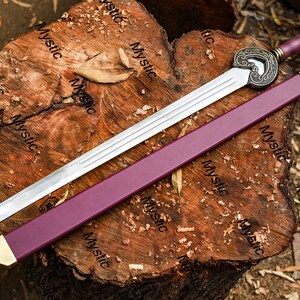 Puede incluir: Una espada larga y plateada con empu&ntilde;adura y vaina burdeos, apoyada sobre una superficie de madera texturizada. La espada presenta una guarda ornamentada de color bronce y una punta dorada en la vaina. El mango tiene un extremo decorativo.