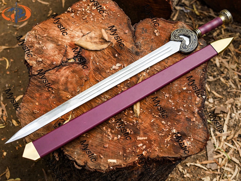 Puede incluir: Una espada larga y plateada con empu&ntilde;adura y vaina burdeos, apoyada sobre una superficie de madera texturizada. La espada presenta una guarda ornamentada de color bronce y una punta dorada en la vaina. El mango tiene un extremo decorativo.