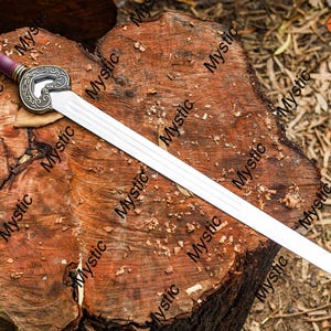 Puede incluir: Una espada decorativa con una hoja plateada y un mango con un pomo dorado y un agarre burdeos. La espada descansa sobre una superficie de madera marr&oacute;n desgastada. La guarda de la espada tiene un dise&ntilde;o ornamentado.