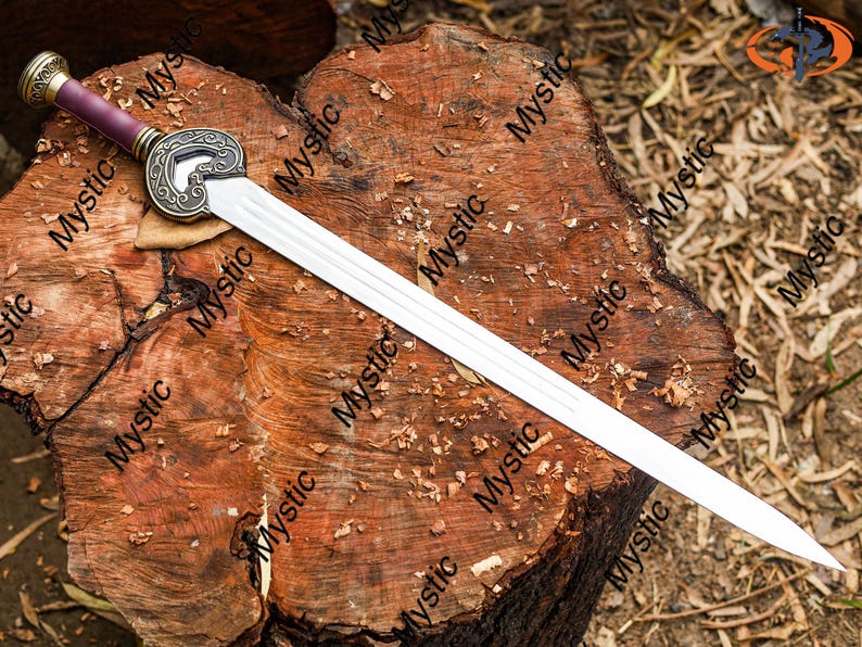 Puede incluir: Una espada decorativa con una hoja plateada y un mango con un pomo dorado y un agarre burdeos. La espada descansa sobre una superficie de madera marr&oacute;n desgastada. La guarda de la espada tiene un dise&ntilde;o ornamentado.