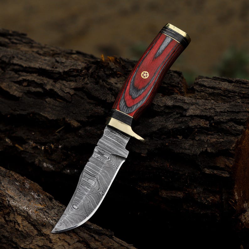 Puede incluir: Un cuchillo de caza de acero de Damasco con un mango de madera roja y negra. El mango tiene detalles de lat&oacute;n y un pasador decorativo. La hoja tiene un borde curvo y un patr&oacute;n distintivo.