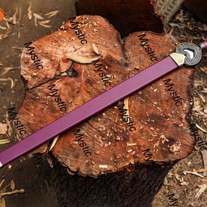 Puede incluir: Una espada morada con una hoja y empu&ntilde;adura doradas, apoyada sobre una superficie de madera texturizada. La espada presenta una guarda decorativa con un dise&ntilde;o en forma de coraz&oacute;n. El mango est&aacute; envuelto en material morado a juego.