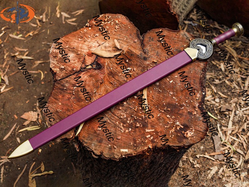 Puede incluir: Una espada morada con una hoja y empu&ntilde;adura doradas, apoyada sobre una superficie de madera texturizada. La espada presenta una guarda decorativa con un dise&ntilde;o en forma de coraz&oacute;n. El mango est&aacute; envuelto en material morado a juego.