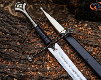 May include: A silver and black sword with a black handle and a silver blade. The sword has a decorative hilt and a black sheath. The blade has an inscription and a star design. The sword is lying on a brown surface.