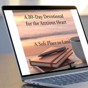 May include: Laptop screen displaying text: "A 30-Day Devotional for the Anxious Heart" and "A Soft Place to Land." The screen shows a stack of brown notebooks, a pen, and a white mug. The background is a blurred beach scene.