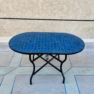 Pode incluir: Mesa de jardim oval com tampo de mosaico azul. A mesa tem uma base de ferro forjado preto com pernas curvas e detalhes decorativos. As dimensões da mesa são aproximadamente 122 cm de comprimento e 76 cm de largura.