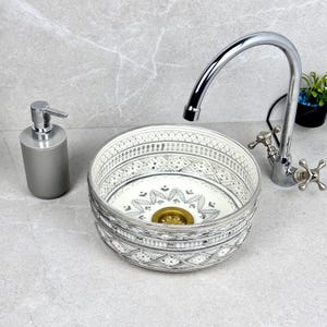May include: A decorative, round bathroom sink with a detailed white and grey pattern. A chrome tap and grey soap dispenser are next to the sink. The sink has a gold-coloured drain.