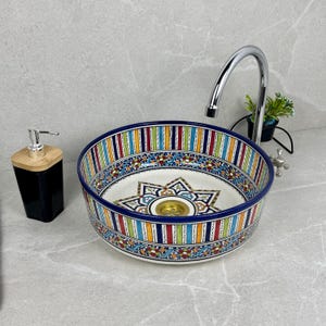 Peut inclure: Un lavabo en céramique blanche avec un motif marocain coloré. Le lavabo a une bordure rayée bleu et blanc avec un motif floral coloré. Le lavabo a un drain en laiton.