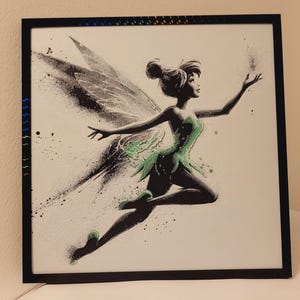 May include: A black framed print of Tinkerbell, a Disney fairy, with green wings and a green dress. The background is white with a splatter effect.