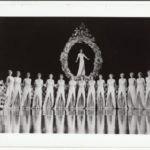 May include: A vintage black and white photograph featuring a group of women in long, flowing dresses with high slits. They stand in a line on a reflective surface, with a woman in a similar dress framed in an ornate circle in the background.