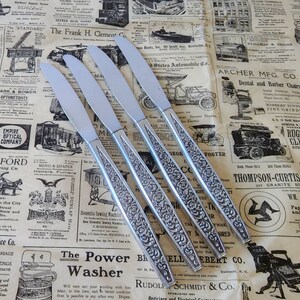 May include: Four stainless steel dinner knives with floral engraved handles. The blades are smooth and shiny. The knives are arranged diagonally on a vintage newspaper background.