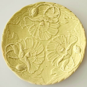 May include: A round, pale yellow ceramic plate with a raised floral design. The plate features three large flowers with detailed petals and dotted centers, surrounded by leaves and buds. The edges of the plate have a slightly rough texture.