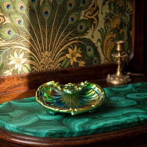 Peut inclure: Un petit plat en forme de coquillage, iris&eacute; de vert et d'or, dot&eacute; d'un bord festonn&eacute; et de trois petits pieds. La surface du plat pr&eacute;sente une finition m&eacute;tallique brillante qui refl&egrave;te la lumi&egrave;re, cr&eacute;ant un effet scintillant sur sa forme.