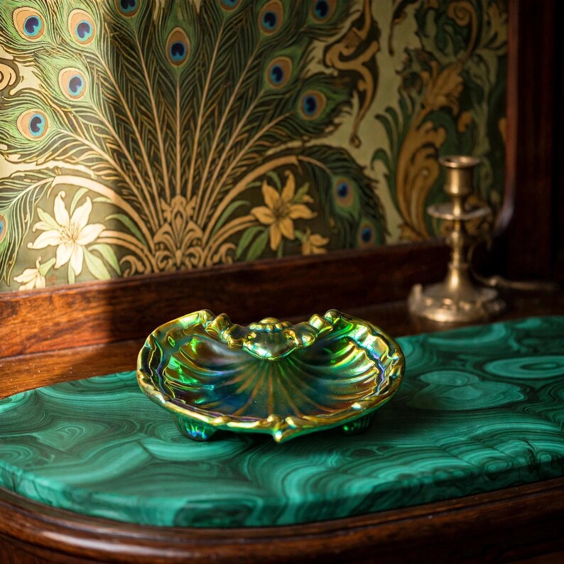 Peut inclure: Un petit plat en forme de coquillage, iris&eacute; de vert et d'or, dot&eacute; d'un bord festonn&eacute; et de trois petits pieds. La surface du plat pr&eacute;sente une finition m&eacute;tallique brillante qui refl&egrave;te la lumi&egrave;re, cr&eacute;ant un effet scintillant sur sa forme.