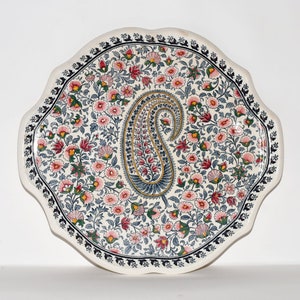 May include: A decorative, scalloped-edge ceramic plate with a floral and paisley design. The plate features a central paisley motif in blue, red, and gold, surrounded by a field of pink and red flowers on a white background. A black border frames the edge.