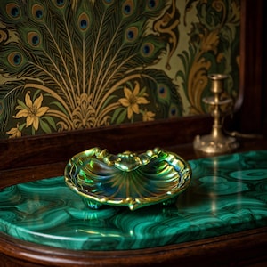 May include: A small, iridescent shell-shaped dish with a scalloped edge and a glossy, multi-colored finish that shifts between green, gold, and blue tones. The dish features a raised, decorative central detail and rests on three small feet.