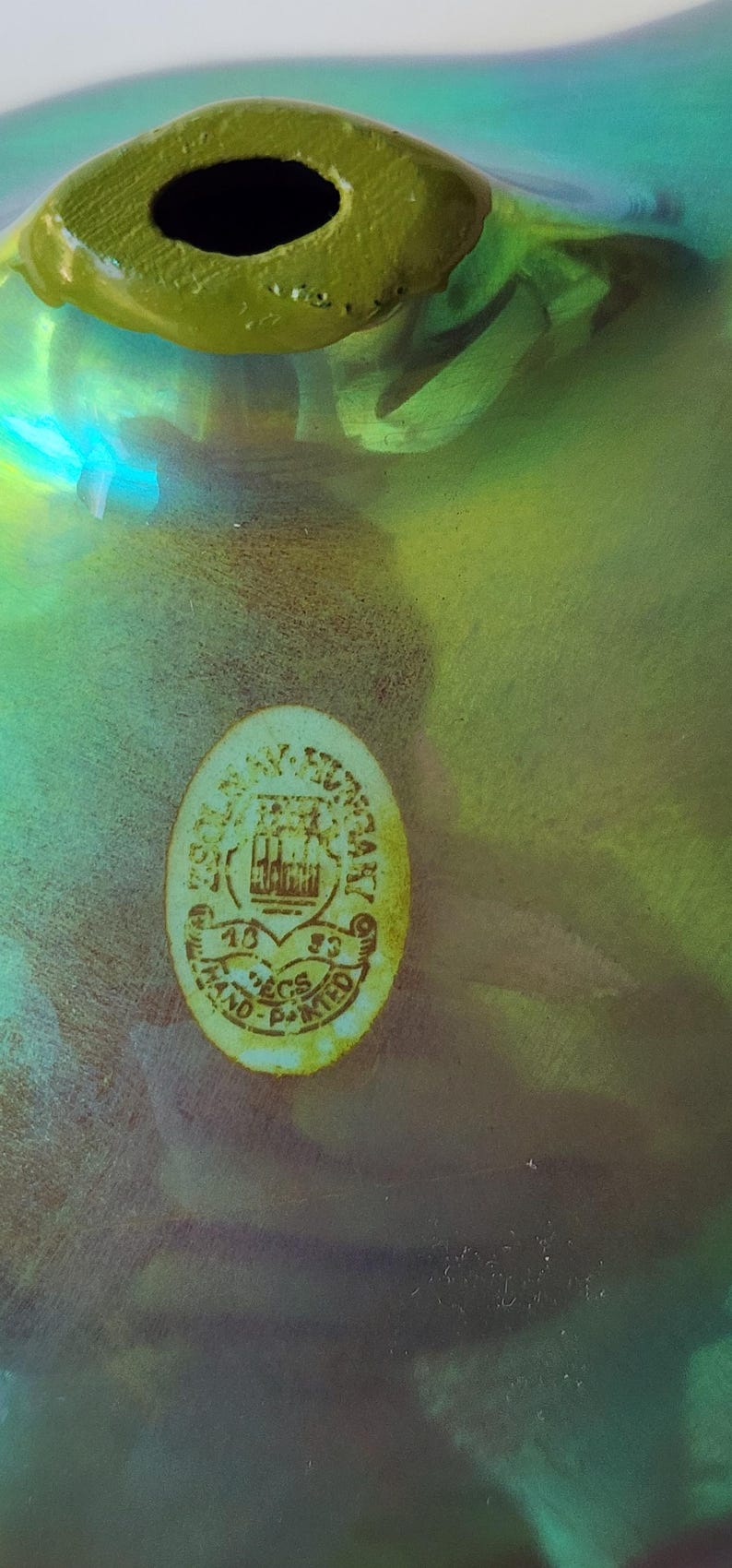 Peut inclure: Un objet en verre vert iris&eacute; avec une ouverture circulaire au sommet. Un autocollant ovale est appos&eacute; sur la surface, portant le texte ZSOLNAY HUNGARY 1853 PECS HAND-PAINTED et une illustration d'armoiries au centre de l'&eacute;tiquette.