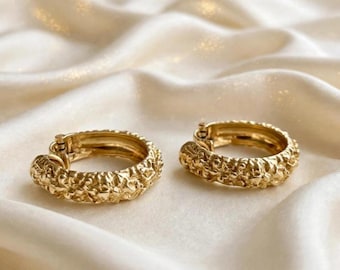Peut inclure: Une paire de créoles dorées présentant une surface texturée et irrégulière. Les boucles d'oreilles sont de forme circulaire avec un mécanisme de fermeture à charnière visible sur le côté intérieur, arborant une finition métallique bosselée.