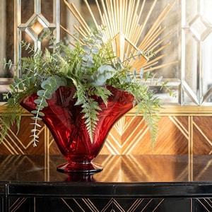 May include: A deep red glass bowl with a wide, ruffled rim and a pedestal base, filled with an arrangement of green eucalyptus branches and fern fronds. The bowl features a smooth, translucent finish and a flared, trumpet-like shape.