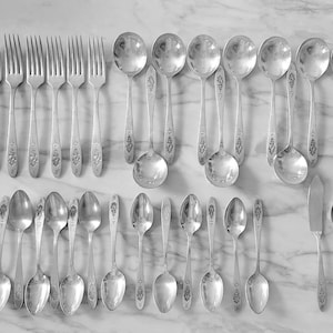 May include: A collection of vintage silverware, including forks, spoons, and a butter knife, arranged on a marble surface. The silverware has a decorative floral pattern on the handles. The spoons are of various sizes, and the set appears to be complete.