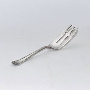 May include: A silver cake fork with a decorative handle and two prongs. The fork has a small design on the top of the handle.