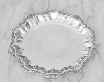 Peut inclure: Un plateau de service argenté poli avec une surface réfléchissante et un motif floral en relief complexe au centre. Le plateau présente un bord surélevé festonné avec des motifs décoratifs en forme de coquille à intervalles réguliers.