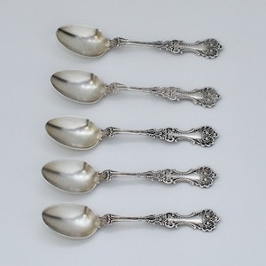 May include: Five silver spoons with ornate, detailed handles and rounded bowls are arranged on a light gray surface. The spoons have a classic, vintage design, with intricate floral and scrollwork patterns on the handles.