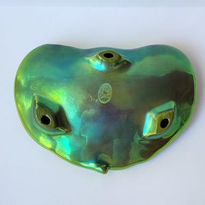 Peut inclure: Un ocarina en c&eacute;ramique aux reflets iris&eacute;s verts et bleus, dot&eacute; d'une forme en c&oelig;ur. L'instrument pr&eacute;sente trois trous de jeu sur la face avant et un petit autocollant ovale pr&egrave;s du centre sup&eacute;rieur. La finition brillante refl&egrave;te la lumi&egrave;re.