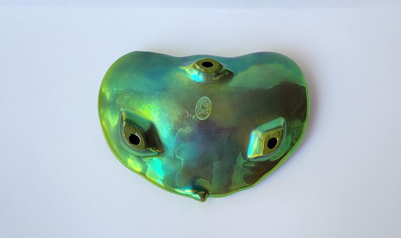 Peut inclure: Un ocarina en c&eacute;ramique aux reflets iris&eacute;s verts et bleus, dot&eacute; d'une forme en c&oelig;ur. L'instrument pr&eacute;sente trois trous de jeu sur la face avant et un petit autocollant ovale pr&egrave;s du centre sup&eacute;rieur. La finition brillante refl&egrave;te la lumi&egrave;re.