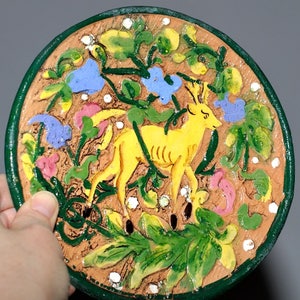 May include: A ceramic plate with a green rim and a yellow deer in a floral design. The deer is surrounded by green leaves, pink and blue flowers, and white dots.