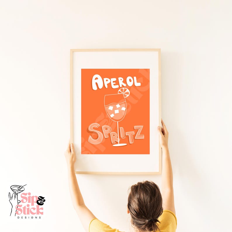 May include: A framed print with an orange background and white text that reads "Aperol Spritz". The print features a white illustration of a glass of Aperol Spritz with an orange slice.