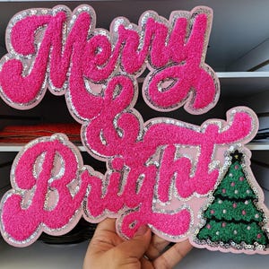 Peut inclure: Un &eacute;cusson rose textur&eacute; avec les mots "Merry & Bright" en &eacute;criture cursive, bord&eacute; de paillettes argent&eacute;es. Un sapin de No&euml;l vert avec des ornements roses et une &eacute;toile est &agrave; droite. Parfait pour les cr&eacute;ations de No&euml;l.