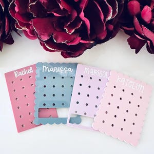 May include: Four pastel-colored earring display cards with scalloped edges and small holes. Each card is labeled with a name: Rachel, Marissa, Marisol, and Katelyn. The background features burgundy flowers.