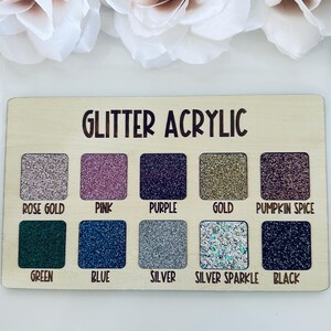 May include: Wooden swatch board with the text "GLITTER ACRYLIC" and ten glitter color samples: rose gold, pink, purple, gold, pumpkin spice, green, blue, silver, silver sparkle, and black. White flowers are in the background.