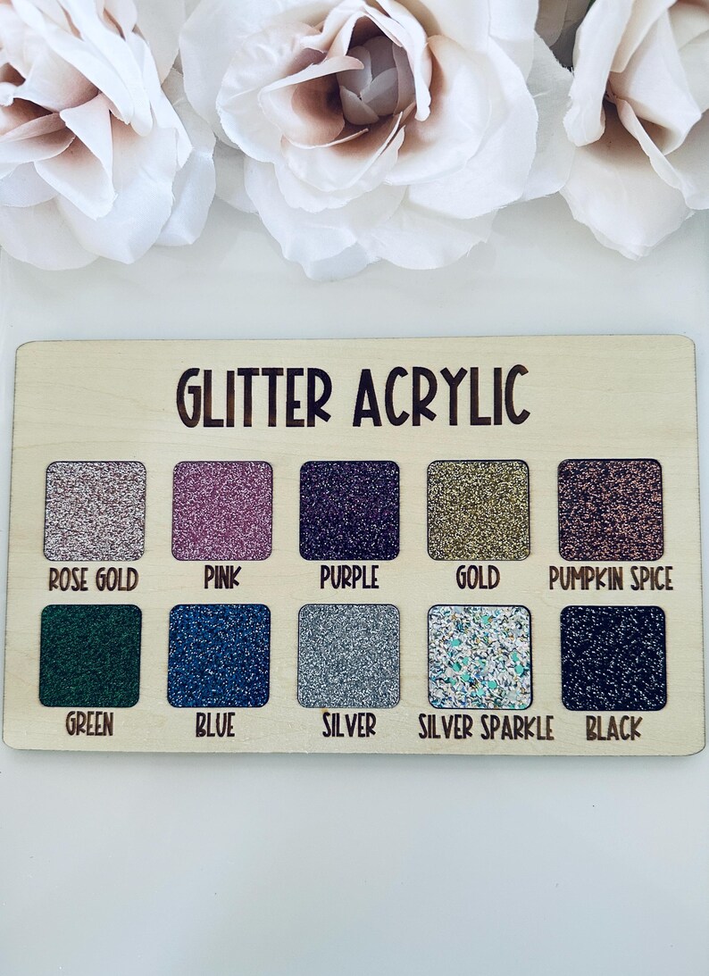 May include: Wooden swatch board with the text "GLITTER ACRYLIC" and ten glitter color samples: rose gold, pink, purple, gold, pumpkin spice, green, blue, silver, silver sparkle, and black. White flowers are in the background.