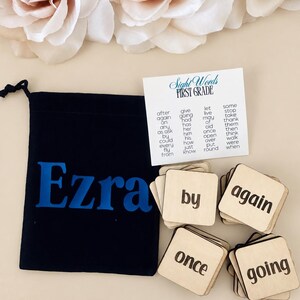 May include: A black drawstring pouch with the name Ezra printed in blue, accompanied by a white card titled Sight Words FIRST GRADE and several square wooden tiles featuring black printed words including by, again, once, and going.