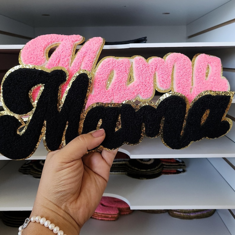 Peut inclure: Un &eacute;cusson rose et noir "Mama" avec une bordure dor&eacute;e &agrave; paillettes. Le mot "Mama" est &eacute;crit dans une police cursive, le mot du haut en rose et le mot du bas en noir. L'&eacute;cusson est tenu devant une &eacute;tag&egrave;re blanche.