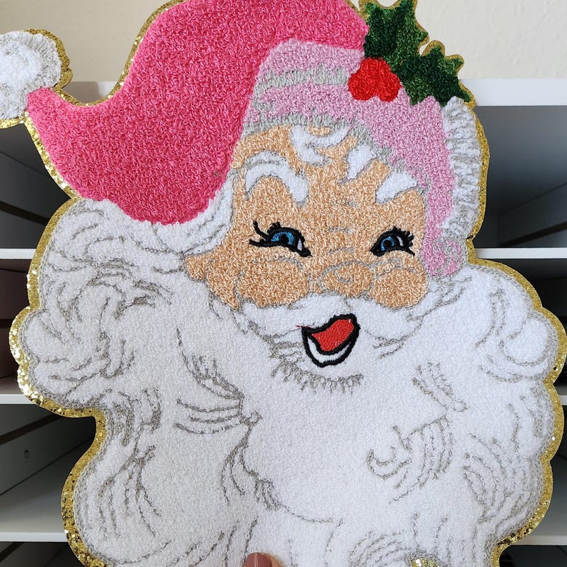 Peut inclure: D&eacute;coration de visage de P&egrave;re No&euml;l avec un bonnet rose, une barbe blanche et une bordure dor&eacute;e scintillante. Le P&egrave;re No&euml;l a les yeux bleus, une bouche rouge et un teint clair. Le design est fait d'un mat&eacute;riau textur&eacute;, parfait pour les f&ecirc;tes.