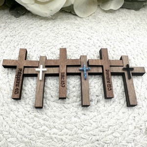 May include: A collection of wooden crosses with varying designs. Some crosses have text, including "Philippians 4:13", "Luke 1:37", and "Romans 12:2". The crosses feature white, blue glitter, and black accents.