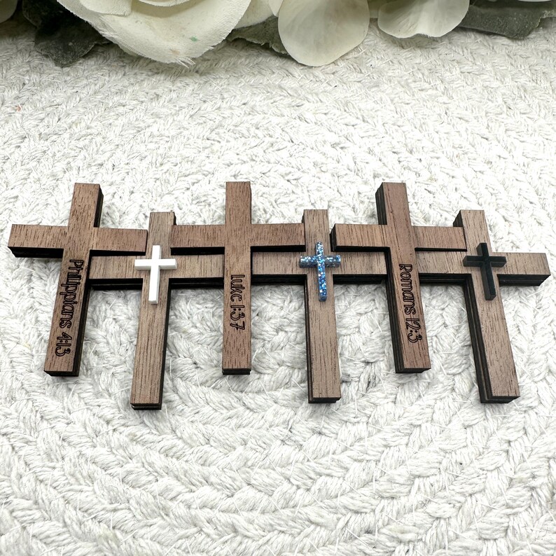 May include: A collection of wooden crosses with varying designs. Some crosses have text, including "Philippians 4:13", "Luke 1:37", and "Romans 12:2". The crosses feature white, blue glitter, and black accents.