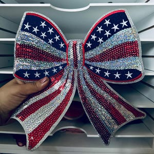 May include: A large decorative bow in the style of the American flag. The bow features a blue border with white stars, red and silver stripes, and is embellished with sequins. The bow is held by a hand.