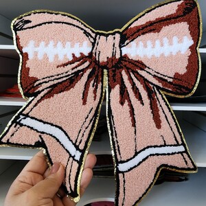 May include: A large, decorative bow in shades of pink, brown, and white, with a gold metallic trim. The bow has a textured, tufted appearance and a black outline. The bow is a craft item, possibly for decorating or embellishing.