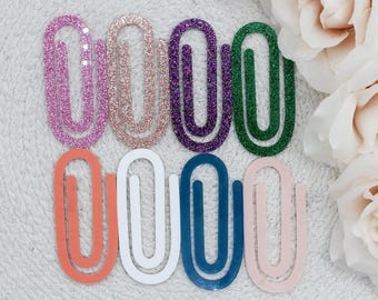 Peut inclure: Une collection de trombones colorés dans diverses nuances, dont rose, or rose, violet, vert, corail, blanc, bleu et rose clair. Certains clips ont des accents de paillettes. Les trombones sont disposés sur une surface blanche texturée.
