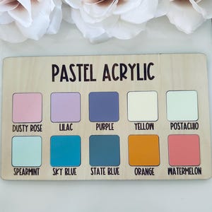 May include: A wooden color palette with the text "PASTEL ACRYLIC" and ten color swatches. Colors include dusty rose, lilac, purple, yellow, pistachio, spearmint, sky blue, state blue, orange, and watermelon. White flowers are in the background.