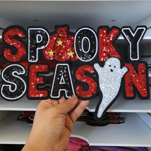 Peut inclure: Un &eacute;cusson sur le th&egrave;me d'Halloween avec les mots "Spooky Season" en paillettes noires et rouges. La lettre "O" est une citrouille avec des &eacute;toiles jaunes, et un fant&ocirc;me blanc est pr&eacute;sent&eacute;. L'&eacute;cusson est tenu par une main.