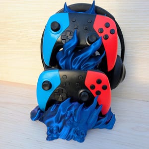 May include: A blue 3D-printed stand featuring sculpted flame and skull motifs, designed to hold two game controllers and a pair of black over-ear headphones. The controllers have black central bodies with bright blue left and red right grips.