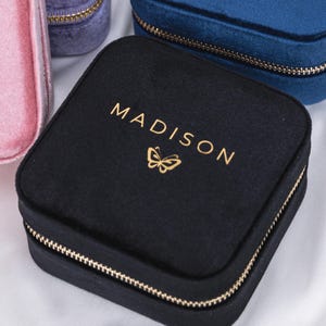 K&ouml;nnte beinhalten: Drei Samt-Schmuckk&auml;stchen mit goldenen Rei&szlig;verschl&uuml;ssen. Die K&auml;stchen sind in Pink, Blau und Schwarz. Das schwarze K&auml;stchen hat einen goldenen Schmetterling und den Text "MADISON" auf der Oberseite. Das blaue K&auml;stchen hat den Text "CHARLIE" und zwei goldene Pfeile auf der Oberseite.