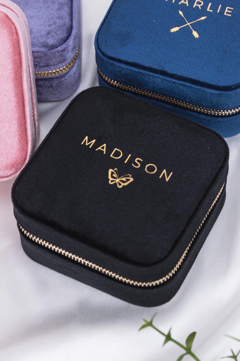 K&ouml;nnte beinhalten: Drei Samt-Schmuckk&auml;stchen mit goldenen Rei&szlig;verschl&uuml;ssen. Die K&auml;stchen sind in Pink, Blau und Schwarz. Das schwarze K&auml;stchen hat einen goldenen Schmetterling und den Text "MADISON" auf der Oberseite. Das blaue K&auml;stchen hat den Text "CHARLIE" und zwei goldene Pfeile auf der Oberseite.
